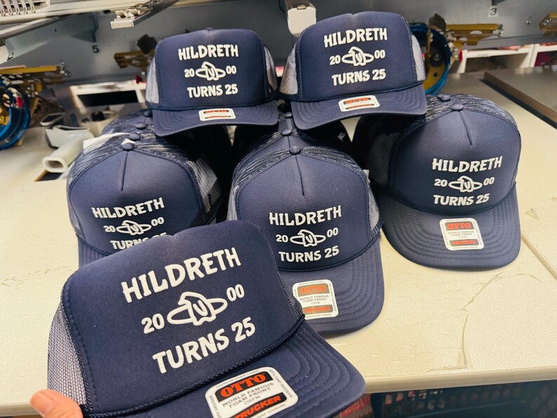 Op de afbeelding: Een collectie marineblauwe truckerpetten met een witte mesh achterkant. Elke pet heeft witte bedrukte tekst met HILDRETH 2000 TURNS 25 en een centrale afbeelding. De petten hebben een gestructureerde schuimrubberen voorkant en een rechthoekige sticker.