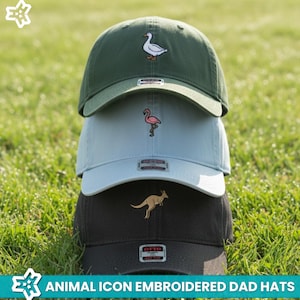 May include: Three dad hats stacked on top of each other, each with an embroidered animal icon. The top hat is green with a white duck, the middle hat is light blue with a pink flamingo, and the bottom hat is dark brown with a kangaroo. The text at the bottom says "ANIMAL ICON EMBROIDERED DAD HATS".