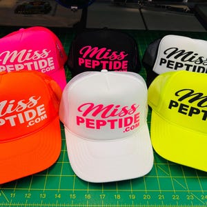 Op de afbeelding: Een collectie van zes truckerpetten in neonoranje, neonroze, zwart, wit en neongeel. Elke pet is voorzien van de tekst "Miss PEPTIDE.com" gedrukt op het voorste schuimrubberen paneel in een contrasterende kleur en heeft een mesh achterkant.