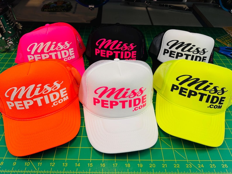 Op de afbeelding: Een collectie van zes truckerpetten in neonoranje, neonroze, zwart, wit en neongeel. Elke pet is voorzien van de tekst "Miss PEPTIDE.com" gedrukt op het voorste schuimrubberen paneel in een contrasterende kleur en heeft een mesh achterkant.