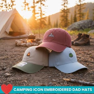 May include: Three embroidered dad hats in burgundy, tan, and light blue, stacked on top of each other. Each hat features a different camping-themed icon: a mountain, a campfire, and a pine tree. The hats are on a bed of dry leaves and twigs.