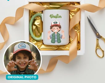 May include: A white ceramic mug with a green handle and rim, featuring a cartoon illustration of a boy named Dustin. The mug is in a white gift box with gold ribbon and a pair of gold scissors.