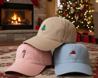 Puede incluir: Tres gorras de béisbol en color beige, rosa y azul claro, cada una bordada con un diseño navideño: un árbol verde, un bastón de caramelo y un gorro de Papá Noel. Las gorras se exhiben frente a un árbol de Navidad y una chimenea.