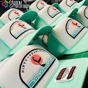 May include: A collection of white and mint green trucker hats with a patch featuring the text "FIFTY + FEARLESS" and "STANIEL CAY 2026". The hats have a mesh back and a curved brim. The hats are labeled "OTTO" and "TRUCKER".