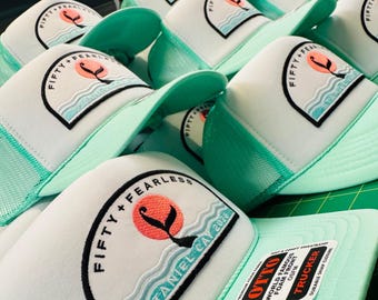 Puede incluir: Una colección de gorras tipo trucker en color verde menta y blanco con un parche circular que muestra un sol coral, la silueta de una cola de ballena negra y olas azules. El texto del parche dice FIFTY + FEARLESS STANIEL CAY 2026.