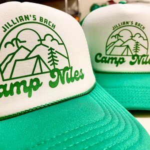 K&ouml;nnte beinhalten: Zwei gr&uuml;ne und wei&szlig;e Trucker-Caps mit dem Text "Jillian's Bach Camp Niles" und einer Grafik mit einem Zelt, Bergen und einem Baum.