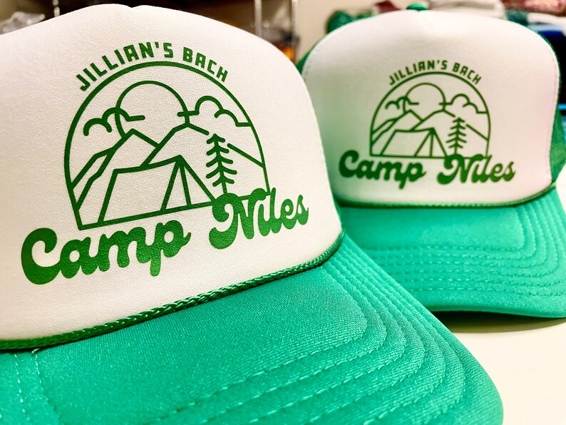 K&ouml;nnte beinhalten: Zwei gr&uuml;ne und wei&szlig;e Trucker-Caps mit dem Text "Jillian's Bach Camp Niles" und einer Grafik mit einem Zelt, Bergen und einem Baum.
