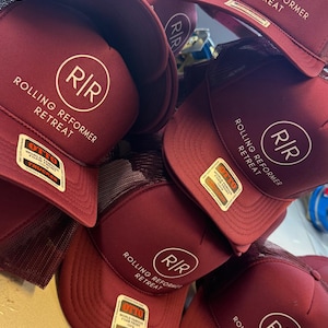 May include: A stack of maroon trucker hats with a white circular logo featuring the letters "R/R" and the words "Rolling Reformer Retreat." Each hat has a mesh back and a curved brim. The hats have a "OTTO" tag.