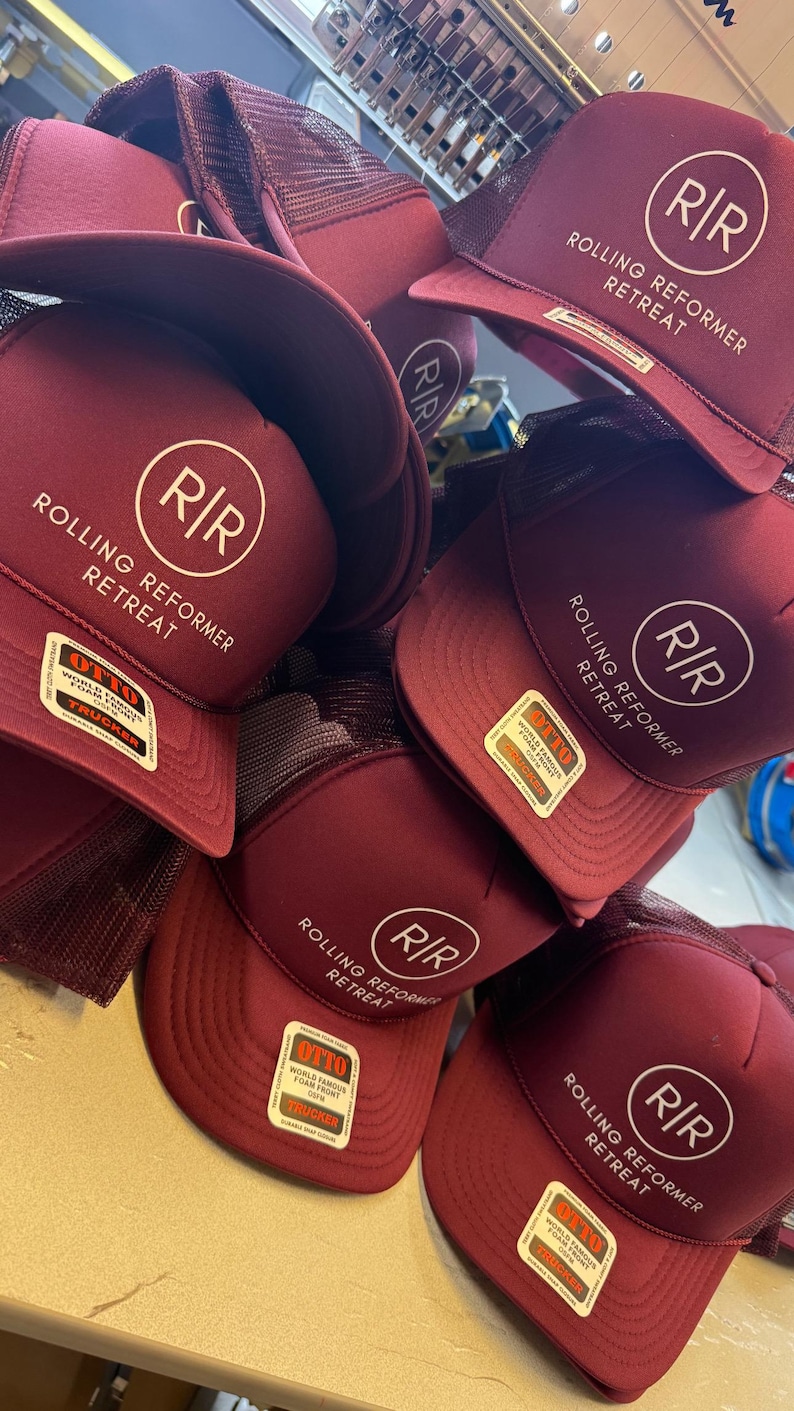 May include: A stack of maroon trucker hats with a white circular logo featuring the letters "R/R" and the words "Rolling Reformer Retreat." Each hat has a mesh back and a curved brim. The hats have a "OTTO" tag.