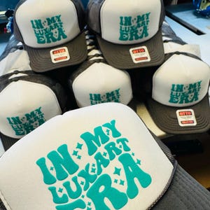 Op de afbeelding: Een collectie antracietgrijze en witte truckerpetten met een schuimrubberen voorpaneel. Elke pet toont de turquoise tekst "IN MY LUXART ERA" met kleine steraccenten. De petten zijn voorzien van een label met de tekst "OTTO".