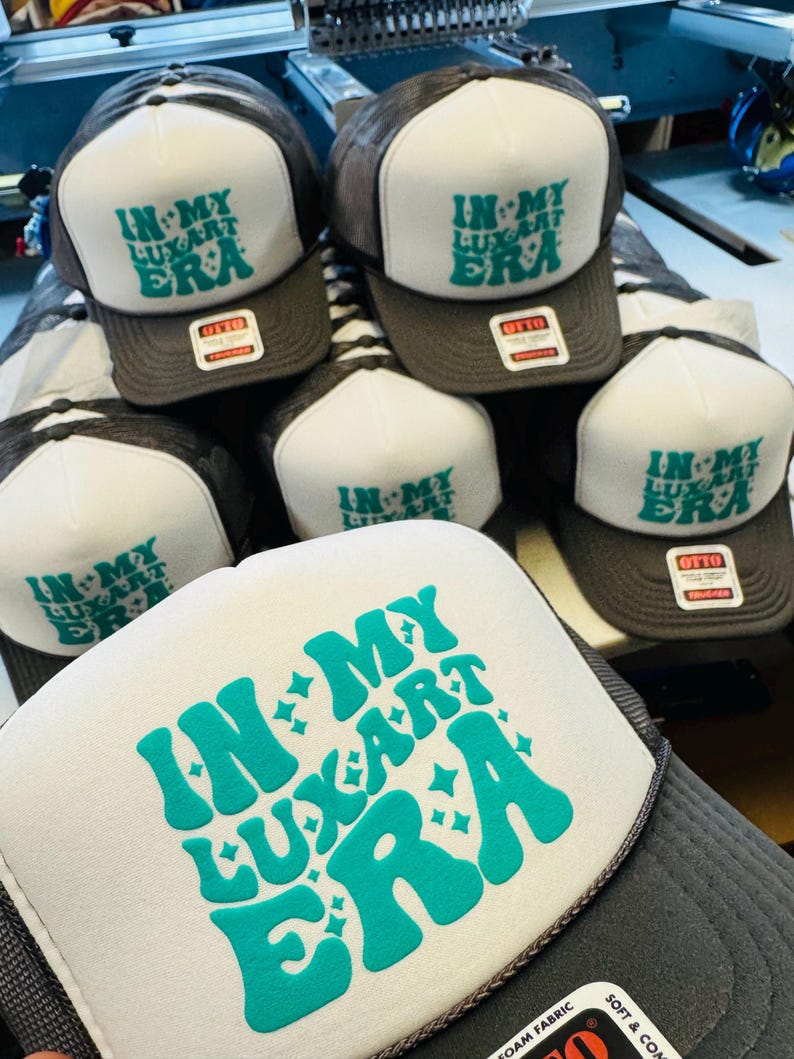 Op de afbeelding: Een collectie antracietgrijze en witte truckerpetten met een schuimrubberen voorpaneel. Elke pet toont de turquoise tekst "IN MY LUXART ERA" met kleine steraccenten. De petten zijn voorzien van een label met de tekst "OTTO".