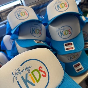 May include: A stack of blue and white trucker hats. Each hat features the text "Northridge KIDS" in a colorful circular design. The hats have a blue mesh back and a white foam front.