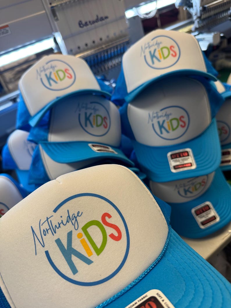 May include: A stack of blue and white trucker hats. Each hat features the text "Northridge KIDS" in a colorful circular design. The hats have a blue mesh back and a white foam front.