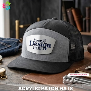 May include: A black and gray acrylic patch hat with a mesh back. The hat features a gray front panel with the text "Your Design HERE" in navy blue. The hat is displayed on a wooden surface.