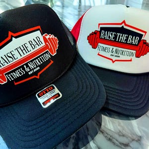 DTF printed OTTO® foam trucker hats with "Raise the Bar Fitness & Nutrition" design. Black and white/red colorways with bold barbell logo, perfect for gym merch or personal trainers. Durable foam front, breathable mesh back.
