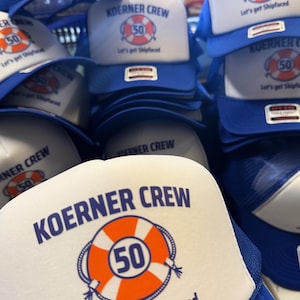 May include: A stack of blue and white trucker hats. The hats have a white front panel with the text "KOERNER CREW" above an orange life preserver with the number "50" inside. Below the graphic is the text "Let's get Shipfaced."