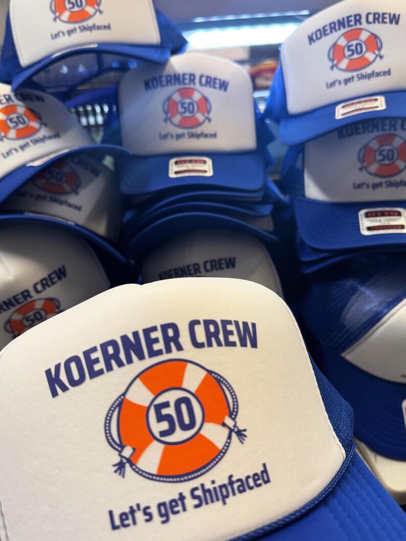 May include: A stack of blue and white trucker hats. The hats have a white front panel with the text "KOERNER CREW" above an orange life preserver with the number "50" inside. Below the graphic is the text "Let's get Shipfaced."