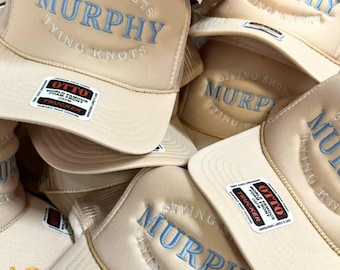 May include: A collection of beige trucker hats featuring light blue embroidered text that reads GIVING SHOTS MURPHY TYING KNOTS. Each hat has a foam front panel, a mesh back, and a black and white Otto brand sticker on the brim.
