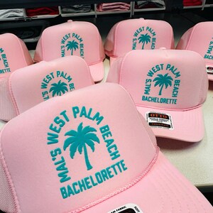 Op de afbeelding: Een collectie lichtroze trucker-petten met een groenblauwe grafische print van een palmboom. De tekst op elke pet luidt Mal's West Palm Beach Bachelorette in een cirkelvormige opstelling rond het palmboomicoon. De petten hebben een schuimrubberen voorkant.