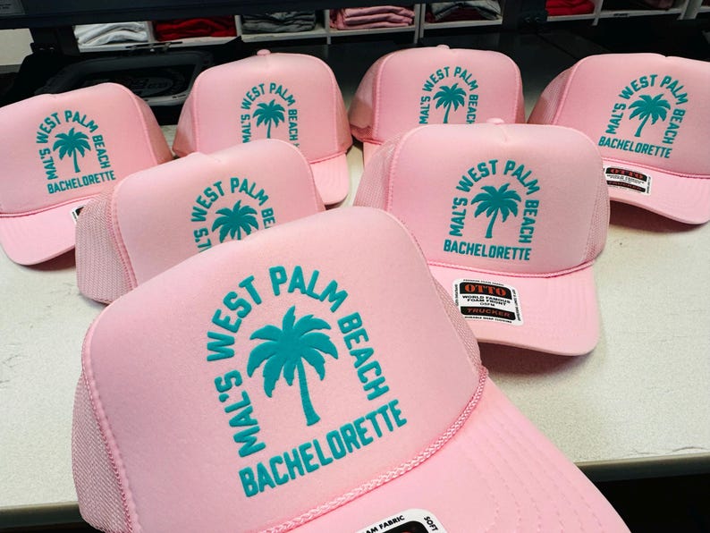 Op de afbeelding: Een collectie lichtroze trucker-petten met een groenblauwe grafische print van een palmboom. De tekst op elke pet luidt Mal's West Palm Beach Bachelorette in een cirkelvormige opstelling rond het palmboomicoon. De petten hebben een schuimrubberen voorkant.