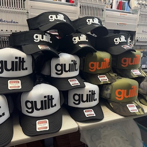 May include: A collection of baseball caps and visors with the word "guilt" in bold white or orange letters. The caps are black, white, and camouflage, with the word "guilt" prominently displayed. Some caps have a mesh back.