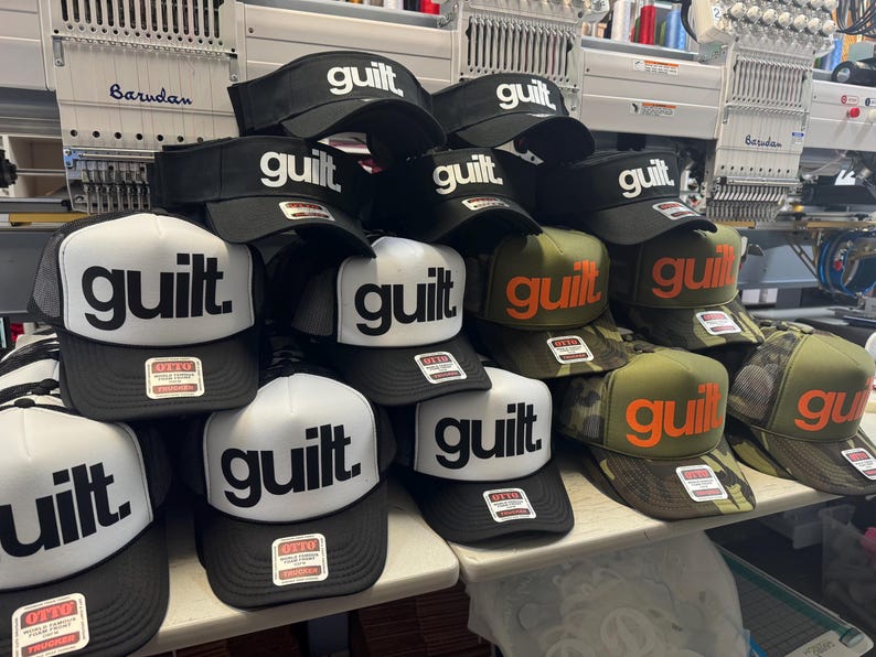 May include: A collection of baseball caps and visors with the word "guilt" in bold white or orange letters. The caps are black, white, and camouflage, with the word "guilt" prominently displayed. Some caps have a mesh back.