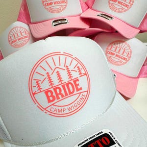 Custom pink and white OTTO foam trucker hats featuring a “Camp Wiggins” design in light red. The bride’s hat is customized with bold “BRIDE” text at the center, while the bridesmaid hats display only “Camp Wiggins.” Perfect for bachelorette parties or group camping trips.