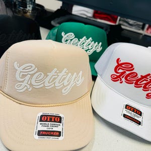 Op de afbeelding: Een collectie van vier trucker-petten met een schuimrubberen voorkant en een mesh achterkant. De petten zijn zwart, groen, beige en wit, elk geborduurd met het woord Gettys in een scriptlettertype. De beige en witte petten hebben een Otto-sticker.