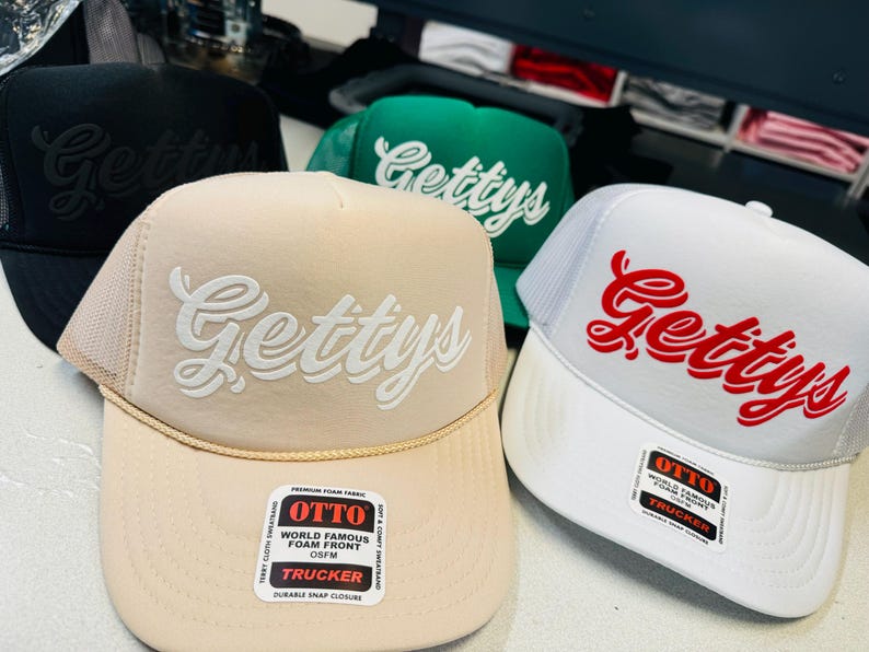 Op de afbeelding: Een collectie van vier trucker-petten met een schuimrubberen voorkant en een mesh achterkant. De petten zijn zwart, groen, beige en wit, elk geborduurd met het woord Gettys in een scriptlettertype. De beige en witte petten hebben een Otto-sticker.