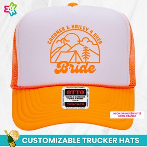 K&ouml;nnte beinhalten: Ein wei&szlig;er und neonoranger Trucker-Hut mit einer schwarzen und orangen Grafik von Bergen, einem Zelt und einer Kiefer. Der Text "GARDNER & HAILEY 4 EVER" und "Bride" ist auf dem Hut aufgedruckt. Der Hut hat einen Druckknopfverschluss und wird von OTTO hergestellt.