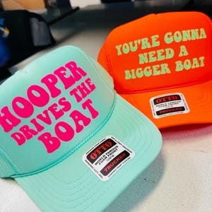 Op de afbeelding: Een mintgroene truckerpet met felroze tekst HOOPER DRIVES THE BOAT en een oranje truckerpet met lichtgroene tekst YOU'RE GONNA NEED A BIGGER BOAT. Beide petten zijn voorzien van Otto-merkstickers op de voorste schuimpanelen.