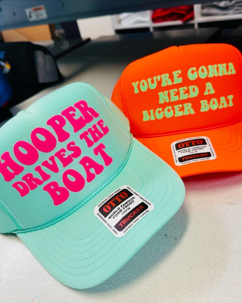 Op de afbeelding: Een mintgroene truckerpet met felroze tekst HOOPER DRIVES THE BOAT en een oranje truckerpet met lichtgroene tekst YOU'RE GONNA NEED A BIGGER BOAT. Beide petten zijn voorzien van Otto-merkstickers op de voorste schuimpanelen.