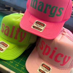 Op de afbeelding: Drie foam trucker petten met mesh achterkant en drukknoopsluiting. De felroze pet heeft de groene tekst "margs", de limoengroene pet heeft roze tekst "salty" en de lichtroze pet toont de rode tekst "spicy". Elke pet is voorzien van een Otto-label.
