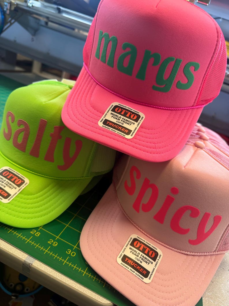 Op de afbeelding: Drie foam trucker petten met mesh achterkant en drukknoopsluiting. De felroze pet heeft de groene tekst "margs", de limoengroene pet heeft roze tekst "salty" en de lichtroze pet toont de rode tekst "spicy". Elke pet is voorzien van een Otto-label.