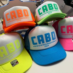 Colorful puff print 'CABO' foam trucker hats with #MYBIGFATRESIDENCYFAMILY 2024 below the main text. Hats come in combinations like neon orange on white, green on white, blue on white, pink on white, and neon yellow on white, displayed on a tabletop