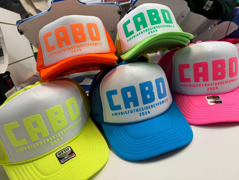 Colorful puff print 'CABO' foam trucker hats with #MYBIGFATRESIDENCYFAMILY 2024 below the main text. Hats come in combinations like neon orange on white, green on white, blue on white, pink on white, and neon yellow on white, displayed on a tabletop