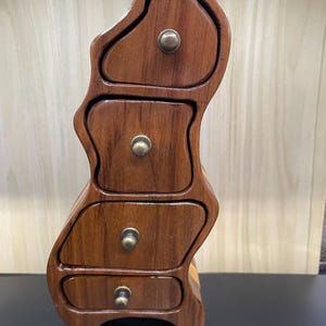May include: A wooden box with four drawers, each with a brass knob. The box is shaped like a wavy, abstract figure and is made of dark brown wood.