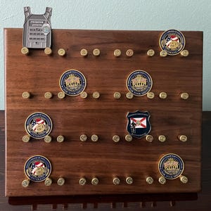 May include: A wooden display board with a silver police officer badge and several round, gold-colored challenge coins. The coins feature various designs, including a flag, a group of people, and a military emblem. The board is decorated with small, gold-colored bullet casings.