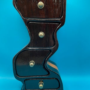 May include: A dark brown wooden jewelry box with four drawers and brass knobs. The box has a unique, curved design and a glossy finish. The drawers are of varying sizes, and the box is set against a blue background.