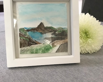 May include: A framed watercolor painting of a landscape with a mountain in the background and a body of water in the foreground. The painting is done in shades of blue, green, and brown.
