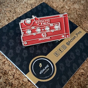 Puede incluir: Un pin rojo Honda DOHC VTEC con forma de cinta de cassette. El pin tiene un borde plateado y el logotipo de Honda. El pin está sobre una tarjeta negra y dorada con el logotipo de Action_Pins e iconos de redes sociales.