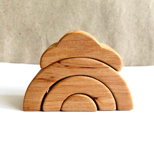 May include: A wooden stacking toy featuring a cloud and rainbow design. The natural wood toy has a cloud-shaped piece on top, with three arched rainbow pieces below. The toy is designed for children.