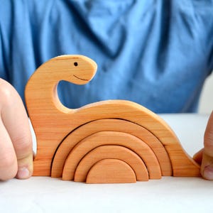Puede incluir: Un juguete de madera con forma de dinosaurio con una cara sonriente. El juguete está hecho de cinco piezas que se encajan y se pueden apilar unas sobre otras.