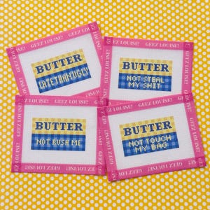 May include: Four rectangular needlepoint canvases with pink borders labeled GEEZ LOUISE. Each features a yellow and blue plaid design with the word BUTTER and phrases: LATE THAN UGLY, NOT STEAL MY SHIT, NOT RUSH ME, and NOT TOUCH MY BAG.
