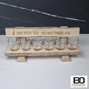 Könnte beinhalten: Holz-Schnapsglas-Halter in Form einer Bank mit dem Text "unser letzter Wille - noch mehr Promille" und zwei gravierten Schnapsgläsern auf der Oberseite. Sechs klare Schnapsgläser sind auf dem Halter platziert.