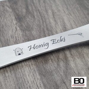 May include: A silver-colored metal honey dipper with the words "Honig Ecki" engraved on it. The dipper features a beehive and a honey dipper illustration. The background is a wooden surface.