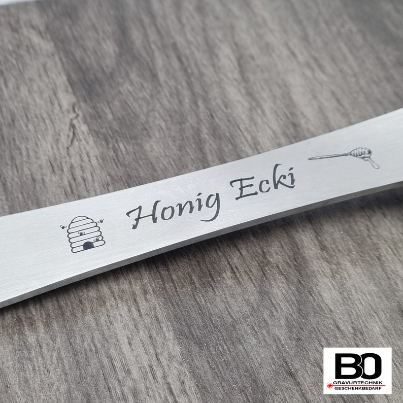 May include: A silver-colored metal honey dipper with the words "Honig Ecki" engraved on it. The dipper features a beehive and a honey dipper illustration. The background is a wooden surface.