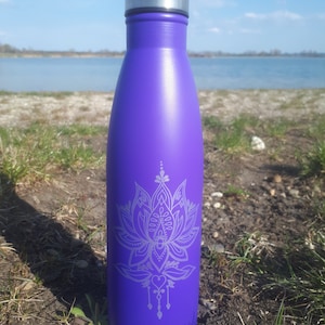 Op de afbeelding: Een paarse roestvrijstalen waterfles met een wit lotusbloemontwerp. De fles heeft een zilveren deksel en staat op een bed van groen gras.