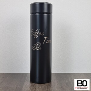 Op de afbeelding: Een zwarte roestvrijstalen reismok met een zwart deksel. De mok heeft de woorden "Coffee Time" in een cursief lettertype, samen met een illustratie van koffiebonen. De mok is cilindrisch en staat rechtop.