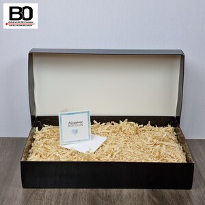 May include: A black gift box with a white interior, filled with wood shavings. A small card with the text "Handgefertigt F&uuml;r Dich" and a blue heart is placed inside the box. The box is open, revealing its contents.